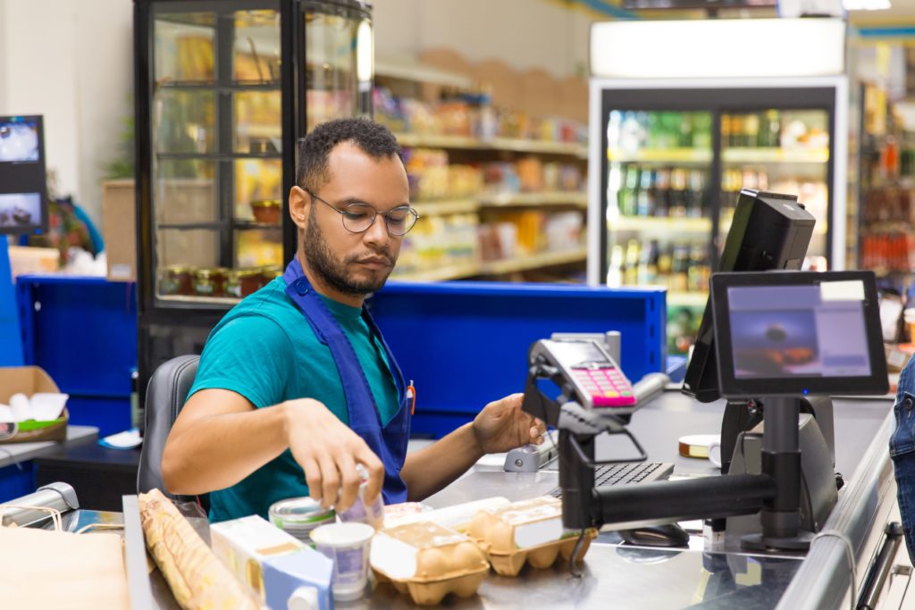 Focused African American cashier scanning goods at checkout. Concentrated young man in eyeglasses at workplace. Shopping concept